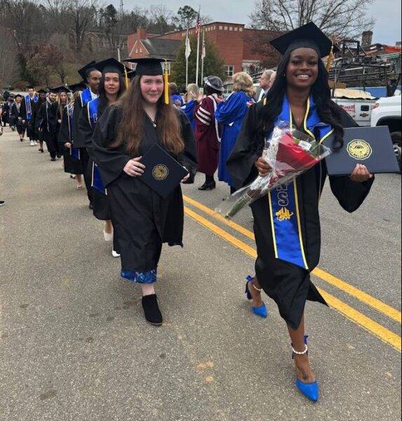 A line of graduates processes up the street, led by a woman wearing blue shoes with her regalia and carrying flowers.