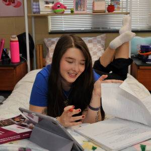 One female student is sitting on her bed with a book and an ipad